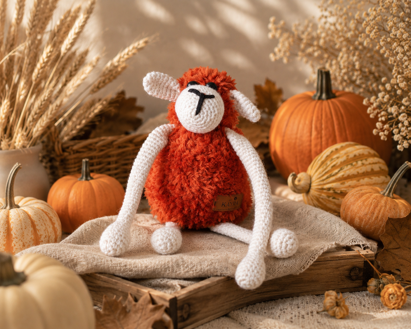 A crocheted plush sheep toy with a fluffy orange-red body, white legs and face, posed on a rustic wooden crate surrounded by pumpkins, dried wheat, and autumn decor.
