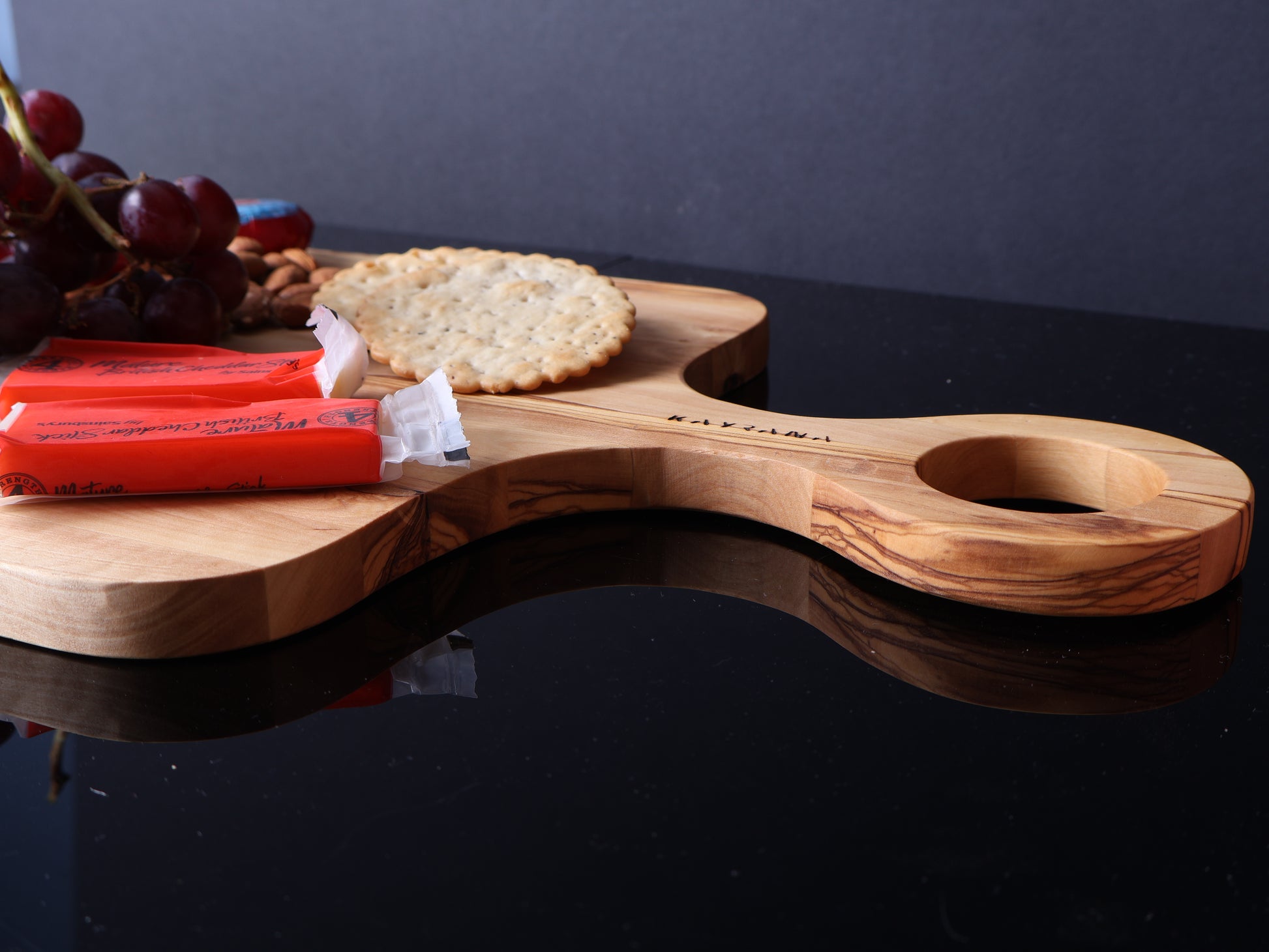 Wooden serving board with a handle, grapes, crackers, and red boxes on a dark surface.