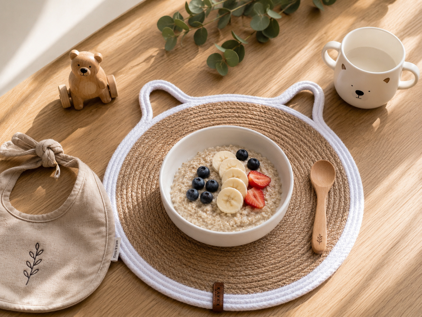 Children's bowl with oatmeal and fruit on a woven mat, next to a bib and cup on a wooden table.