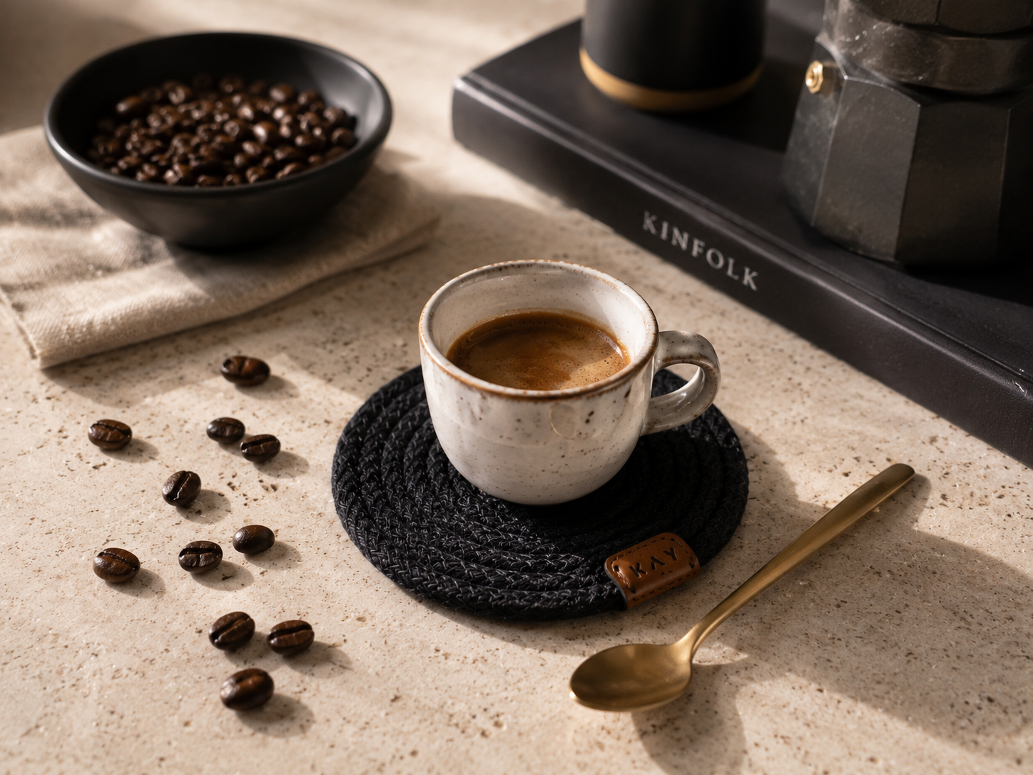 Cup of coffee on a coaster with coffee beans and a spoon on a textured surface, featuring the brand 'KINFOLK'.