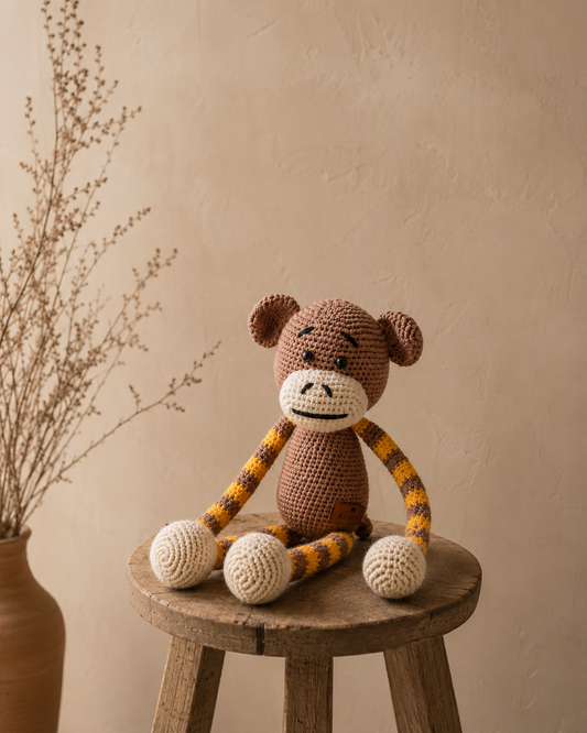 A crocheted monkey plush toy with a brown body, yellow-and-black striped arms and legs, and cream-colored feet and muzzle, sitting on a rustic wooden stool against a warm beige wall with dried branches in a vase.