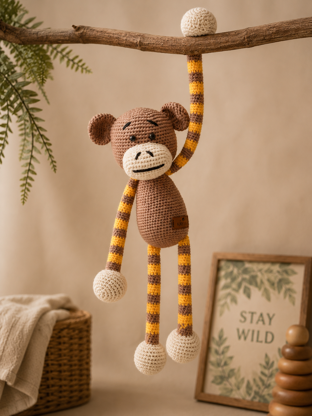 A crocheted monkey hanging from a wooden branch in a cozy room setting, with a basket, towel, and framed 'STAY WILD' sign in the background.