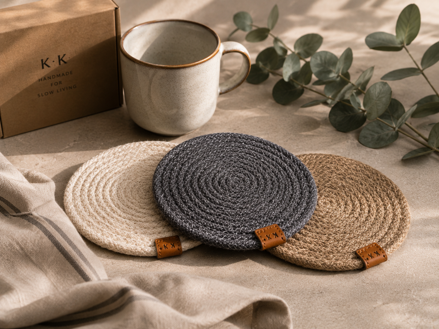 Three round woven coasters in beige, blue, and brown on a surface with a mug and eucalyptus leaves.