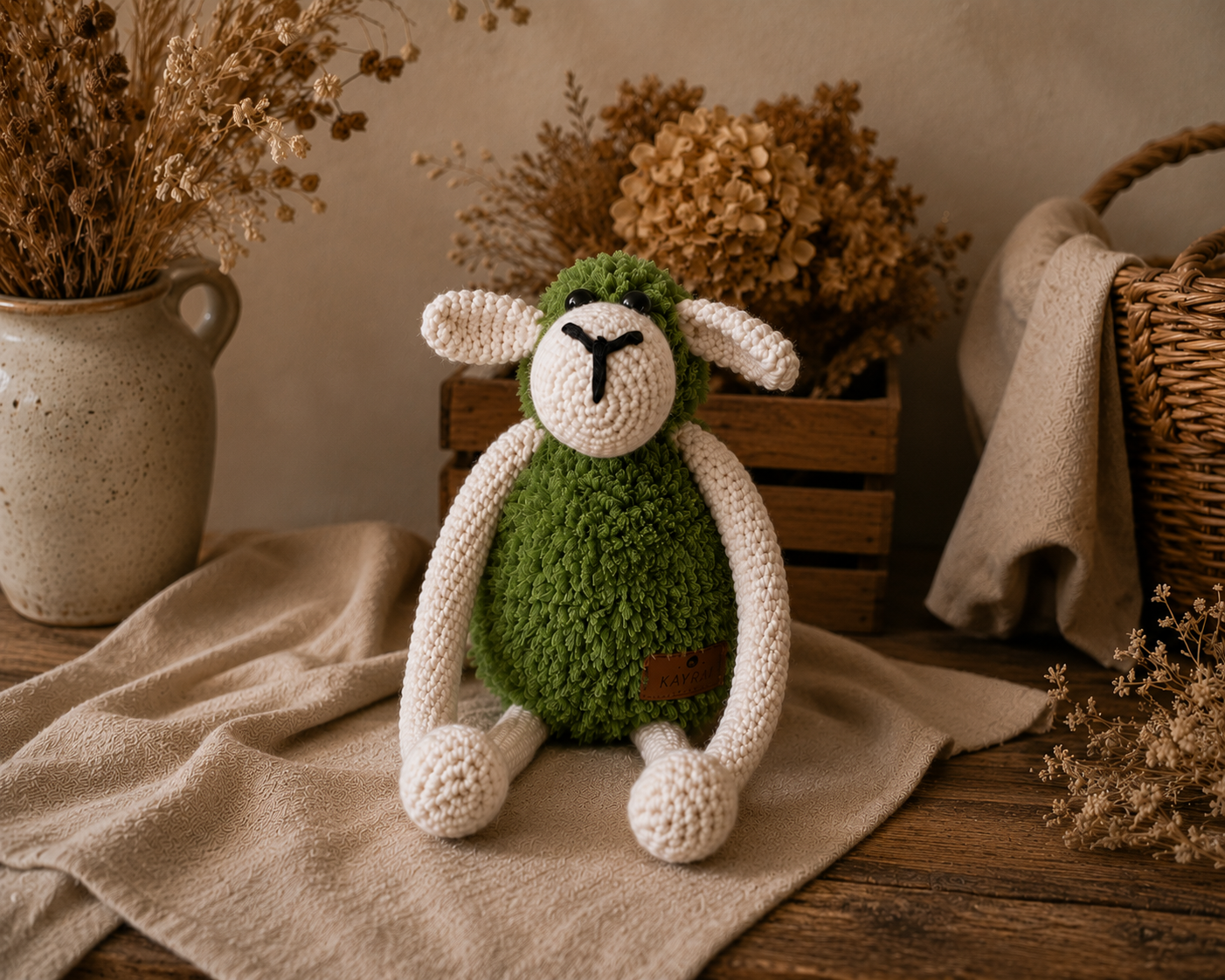 A handmade crocheted plush sheep toy with a green body, white ears and legs, and a black embroidered face, sitting on a beige linen cloth with dried flowers and rustic home decor in the background.