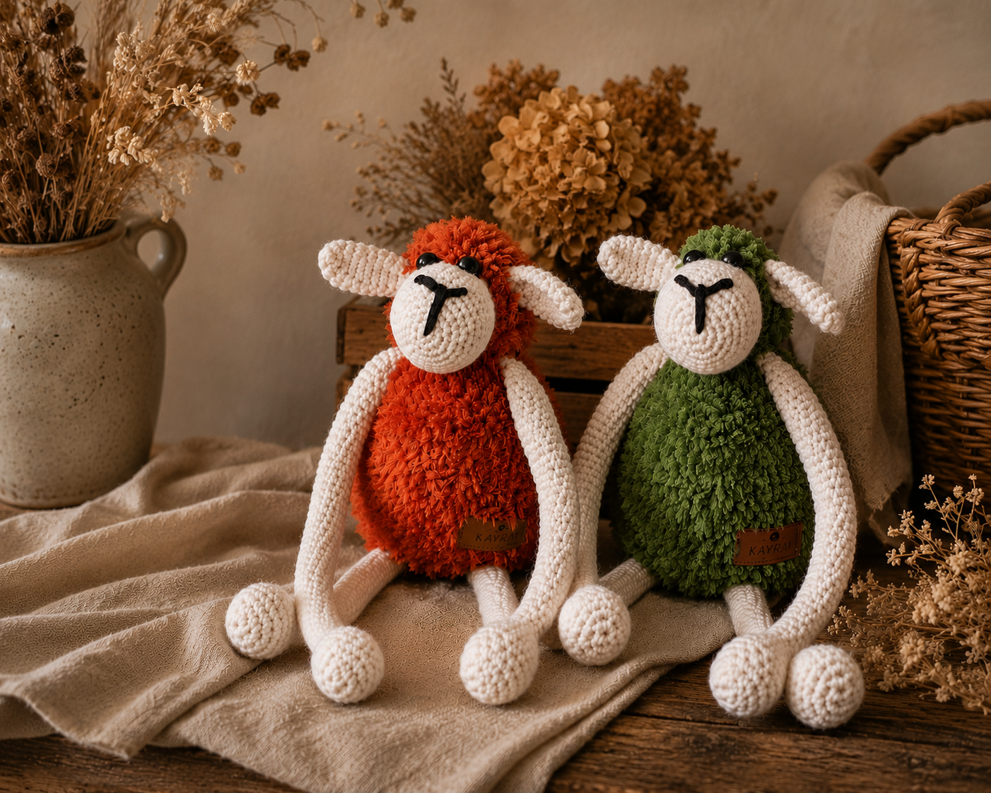 Two crocheted plush sheep toys sitting on a beige cloth in a styled rustic setting with dried flowers, a ceramic vase, and a wicker basket.