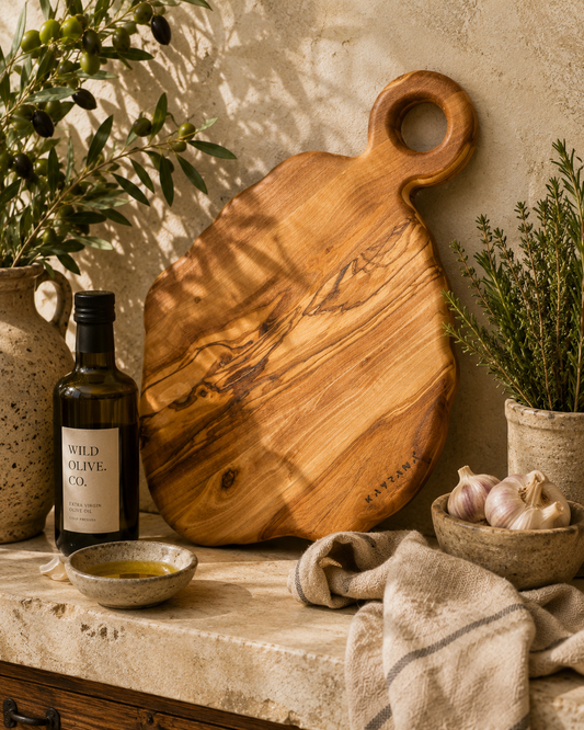 Wooden cutting board on a stone surface with a bottle of Wild Olive Oil, a bowl of olive oil, and garlic.