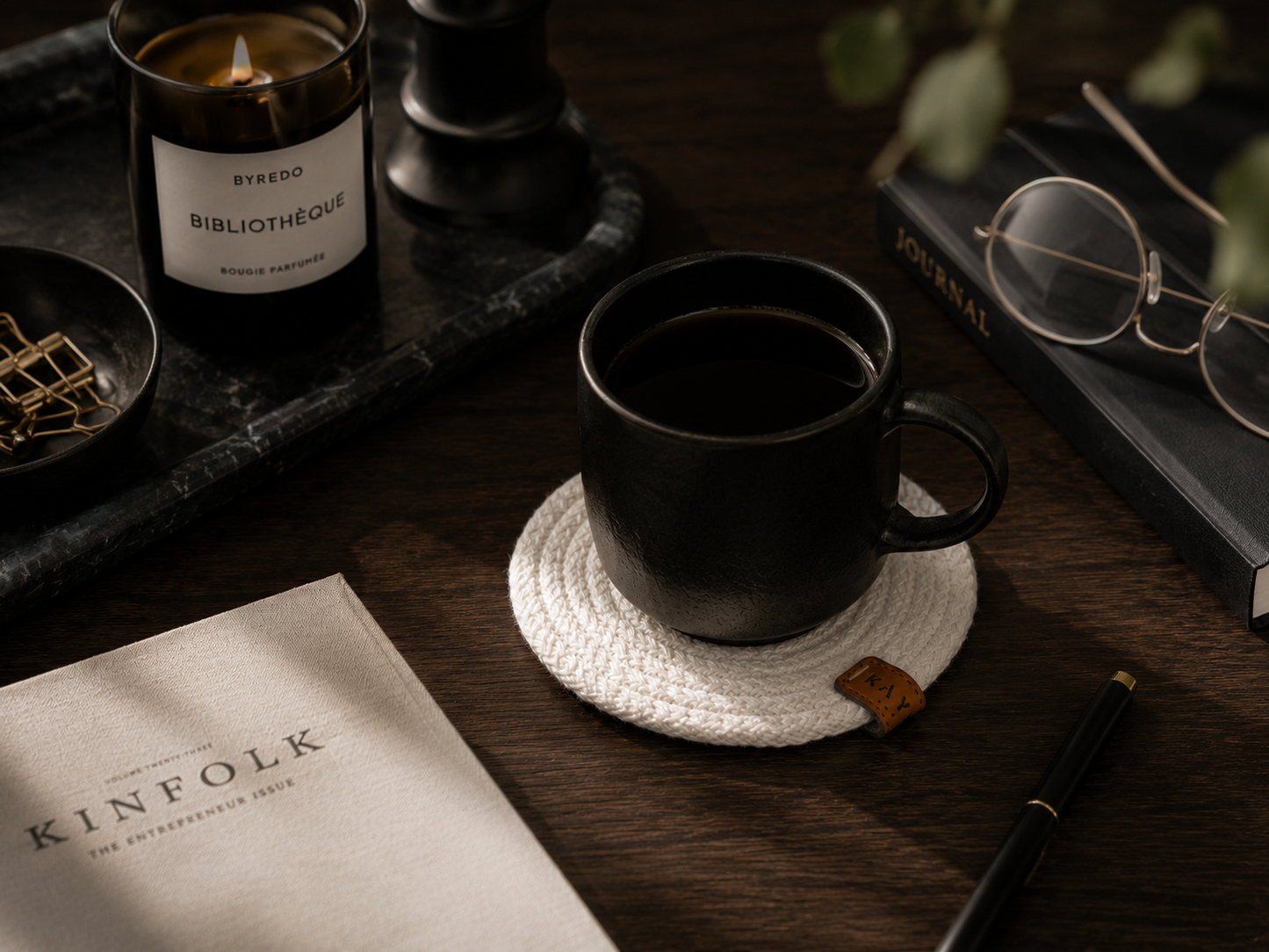 Black mug on a coaster with a candle, book, and glasses on a wooden surface